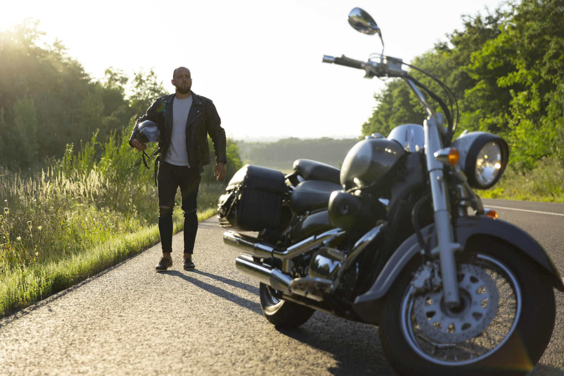 Man walking on a rural roadside holding a motorcycle helmet, cruiser bike parked nearby